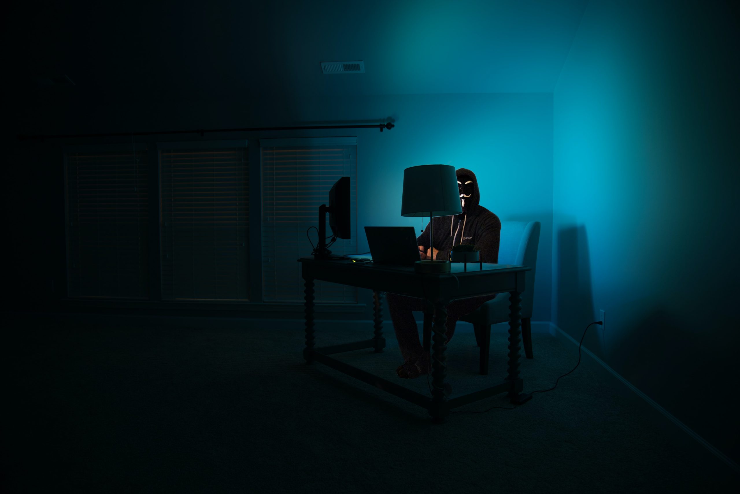 Person in dark room with mask on at a computer hacking.
