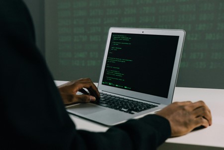 "Stock image showing a person using a laptop at a table, with placeholder command line text in green. We're using this picture to illustrate "cybersecurity".